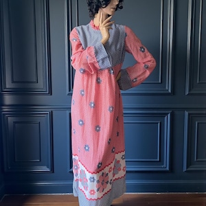 Puede incluir: Un vestido largo de estilo vintage con un estampado de lunares rojos y blancos con detalles florales azules. El vestido tiene mangas largas con puños a cuadros azules y blancos y un corpiño a juego. El dobladillo está decorado con un borde floral.