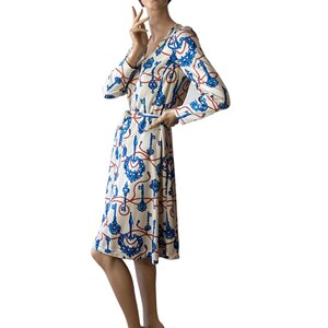 May include: A white dress with a blue and red key pattern. The dress has long sleeves and a V-neckline. It is belted at the waist.