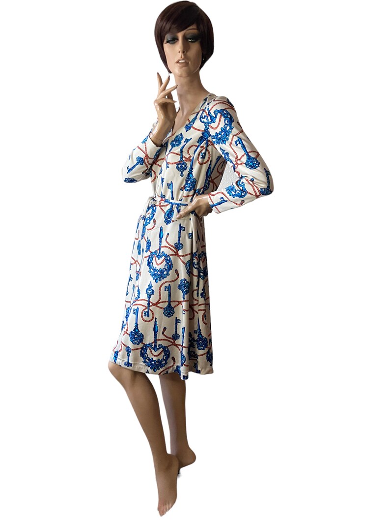 May include: A white dress with a blue and red key pattern. The dress has long sleeves and a V-neckline. It is belted at the waist.