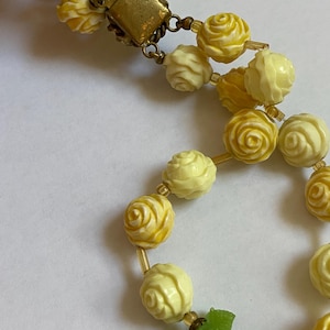 May include: A vintage necklace featuring two strands of carved rose beads in shades of yellow and cream. The necklace has a gold-tone clasp and small, clear beads between the rose beads. Green leaf accents are visible near the clasp.