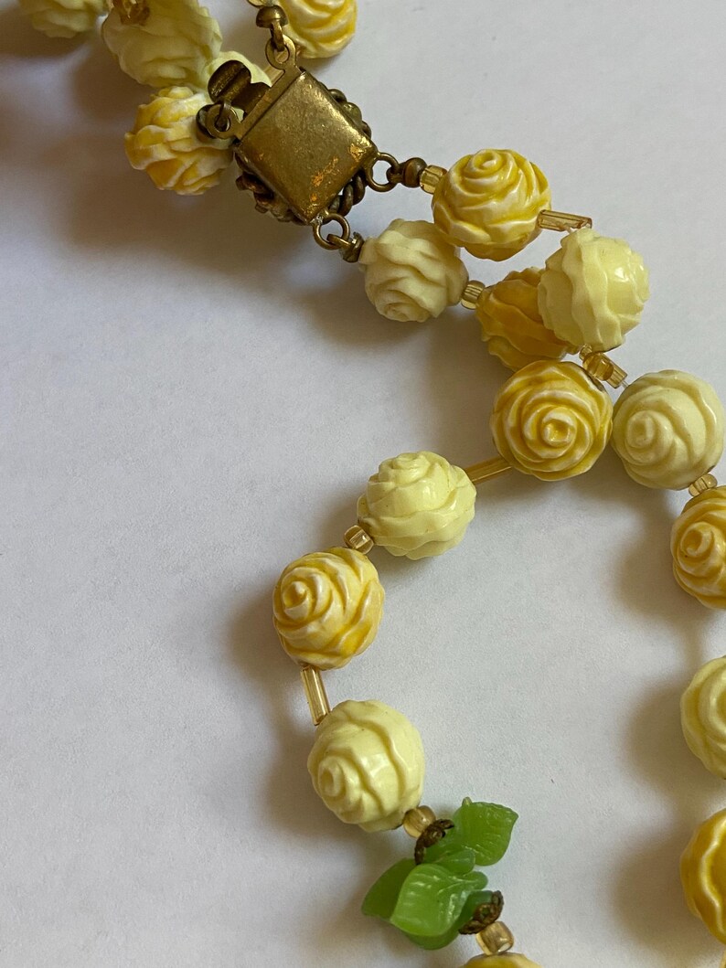 May include: A vintage necklace featuring two strands of carved rose beads in shades of yellow and cream. The necklace has a gold-tone clasp and small, clear beads between the rose beads. Green leaf accents are visible near the clasp.