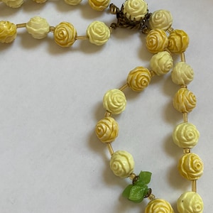 May include: A vintage necklace featuring rose-shaped beads in shades of yellow and cream. The beads are strung together with a delicate gold-colored chain. A small green leaf accent adds a touch of color to the design.