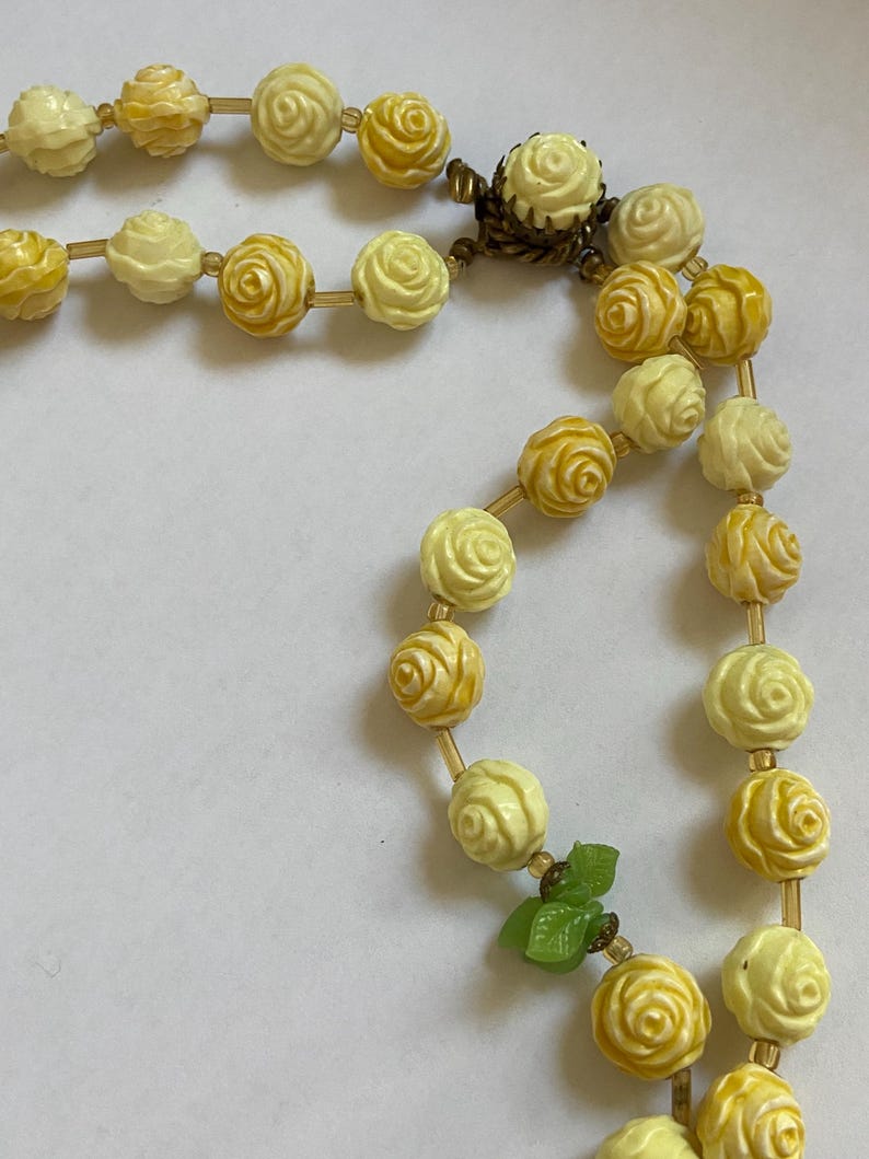 May include: A vintage necklace featuring rose-shaped beads in shades of yellow and cream. The beads are strung together with a delicate gold-colored chain. A small green leaf accent adds a touch of color to the design.
