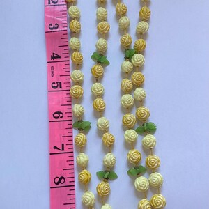 May include: A vintage necklace featuring two strands of cream and yellow rose-shaped beads, accented with green leaf-shaped beads. The necklace is displayed next to a pink measuring tape, showing its length.