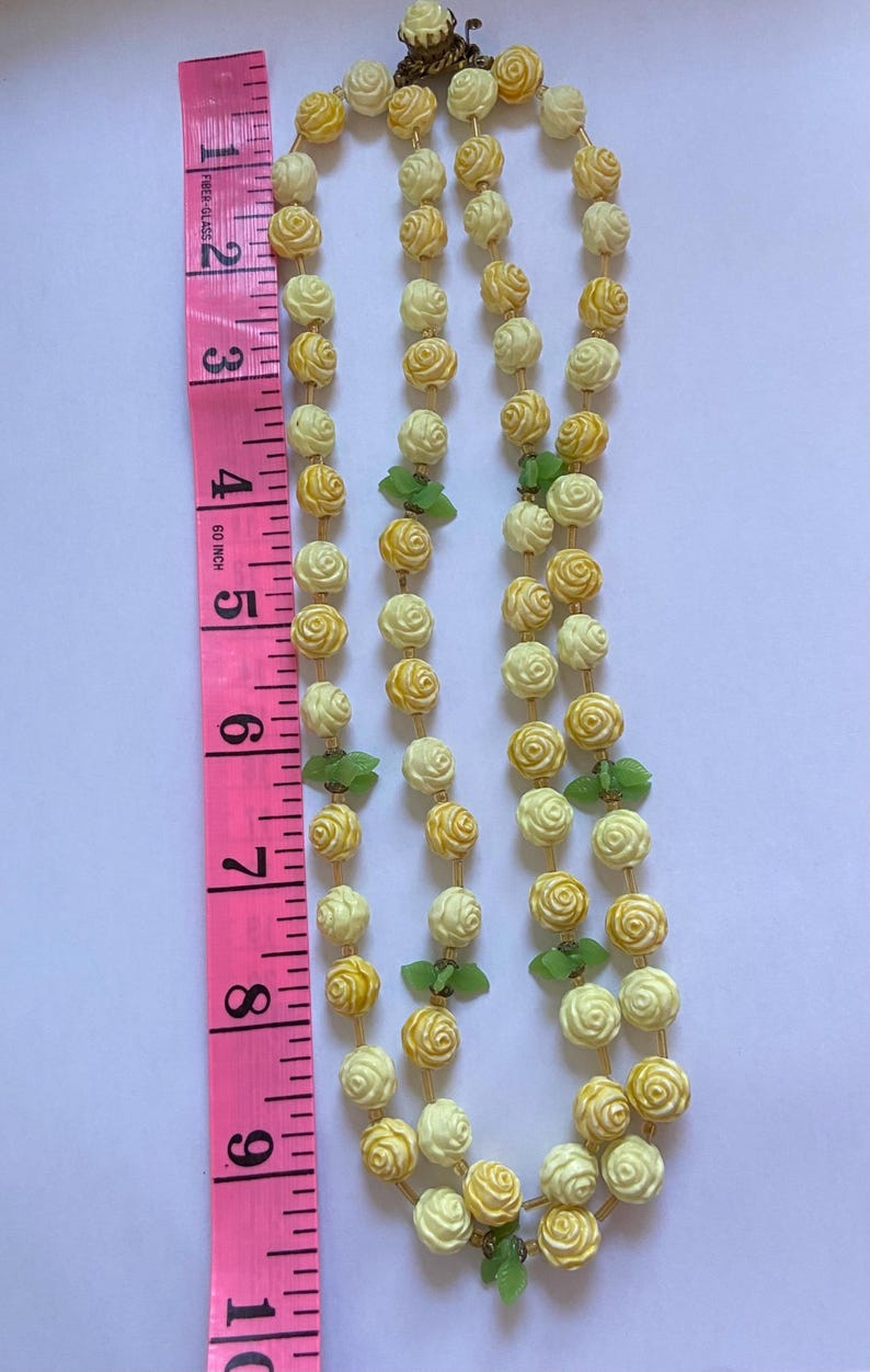 May include: A vintage necklace featuring two strands of cream and yellow rose-shaped beads, accented with green leaf-shaped beads. The necklace is displayed next to a pink measuring tape, showing its length.