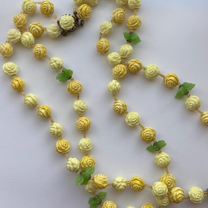 May include: A vintage necklace featuring a double strand of cream and yellow rose-shaped beads. The necklace is accented with small green leaf charms and a decorative clasp. The beads are strung on a gold-colored chain.