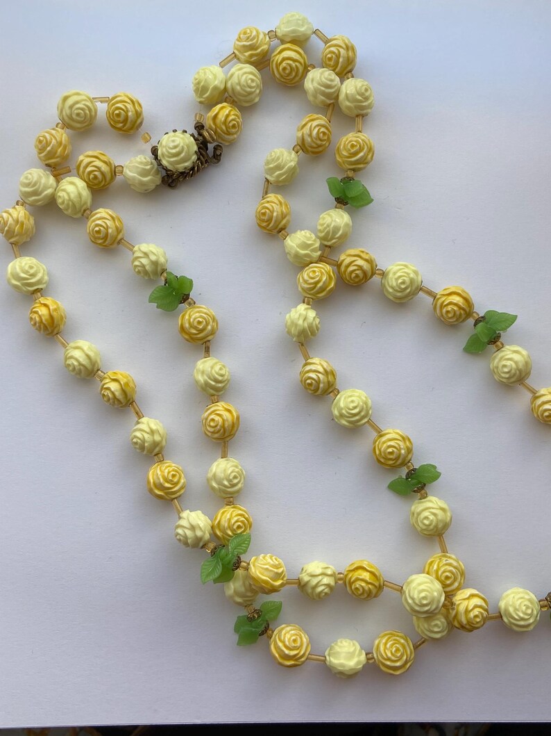May include: A vintage necklace featuring a double strand of cream and yellow rose-shaped beads. The necklace is accented with small green leaf charms and a decorative clasp. The beads are strung on a gold-colored chain.
