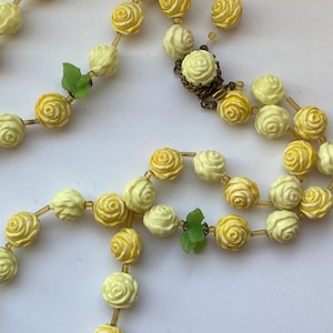 May include: A vintage necklace featuring rose-shaped beads in shades of yellow and cream. The beads are strung together with a delicate chain, and accented with small green leaf details. The necklace has a clasp closure.