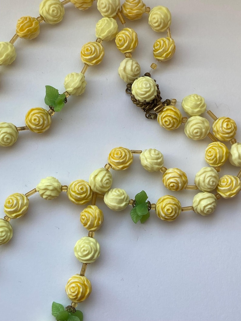 May include: A vintage necklace featuring rose-shaped beads in shades of yellow and cream. The beads are strung together with a delicate chain, and accented with small green leaf details. The necklace has a clasp closure.