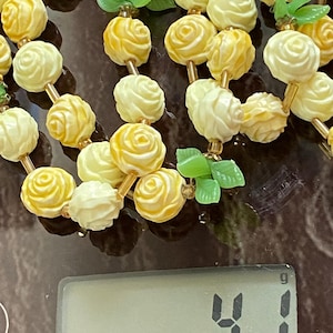 May include: A vintage necklace featuring cream and yellow rose-shaped beads strung together with gold-toned metal. Green leaf accents are interspersed throughout the design. The necklace weighs 41 grams.