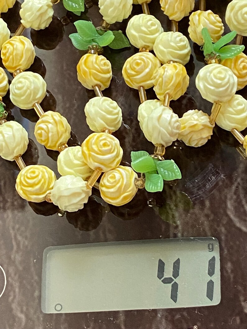 May include: A vintage necklace featuring cream and yellow rose-shaped beads strung together with gold-toned metal. Green leaf accents are interspersed throughout the design. The necklace weighs 41 grams.