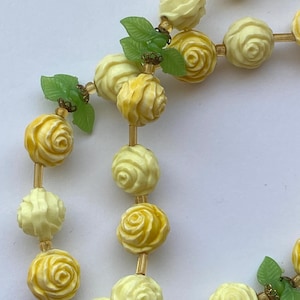 May include: A vintage necklace featuring carved rose beads in shades of yellow and cream. The necklace includes small green leaf accents and is strung on a delicate, gold-toned chain. The rose beads are intricately detailed, showcasing a classic floral design.