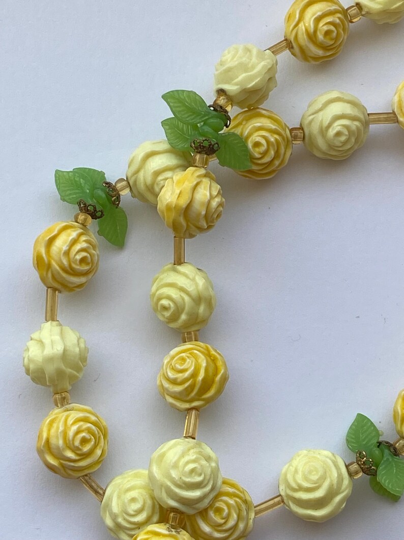 May include: A vintage necklace featuring carved rose beads in shades of yellow and cream. The necklace includes small green leaf accents and is strung on a delicate, gold-toned chain. The rose beads are intricately detailed, showcasing a classic floral design.