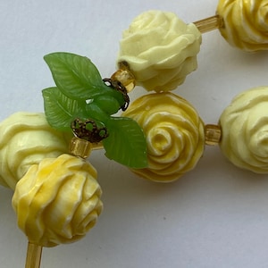 May include: A close-up of a necklace featuring carved rose beads in shades of yellow and cream. The necklace includes green leaf accents and small gold-colored beads strung on a gold-toned chain. The beads are intricately detailed.