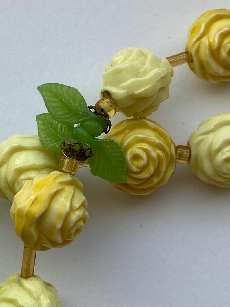 May include: A close-up of a necklace featuring carved rose beads in shades of yellow and cream. The necklace includes green leaf accents and small gold-colored beads strung on a gold-toned chain. The beads are intricately detailed.