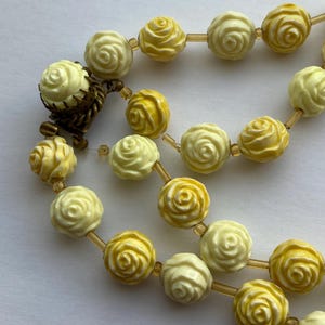 May include: A vintage necklace featuring rose-shaped beads in shades of yellow and cream. The beads are strung together with a delicate, light-brown string. The necklace has a clasp made of a dark, aged metal.