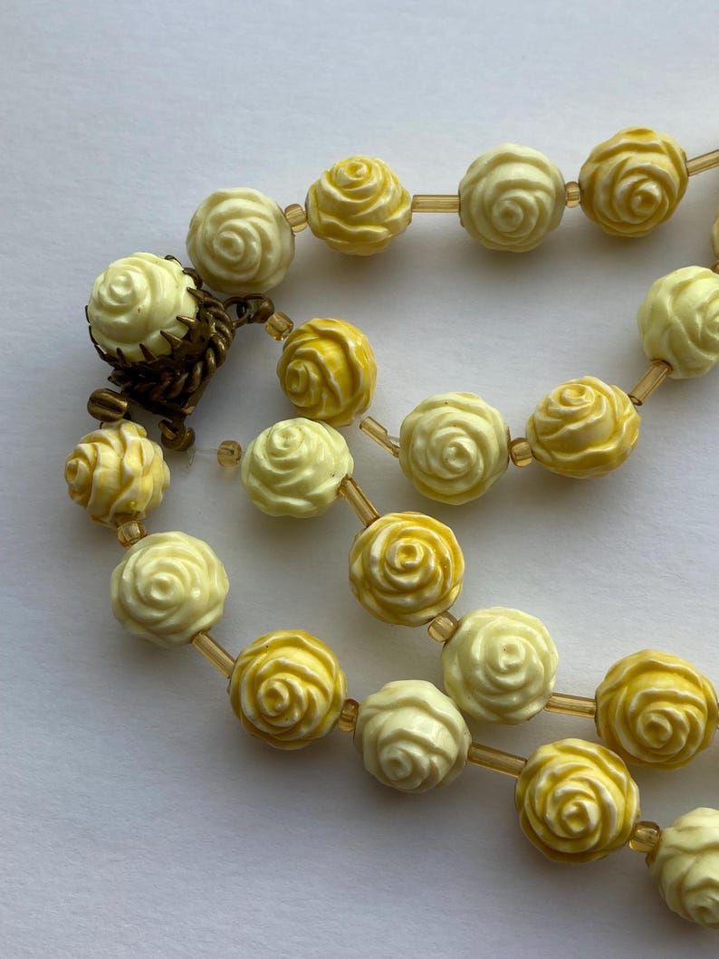May include: A vintage necklace featuring rose-shaped beads in shades of yellow and cream. The beads are strung together with a delicate, light-brown string. The necklace has a clasp made of a dark, aged metal.