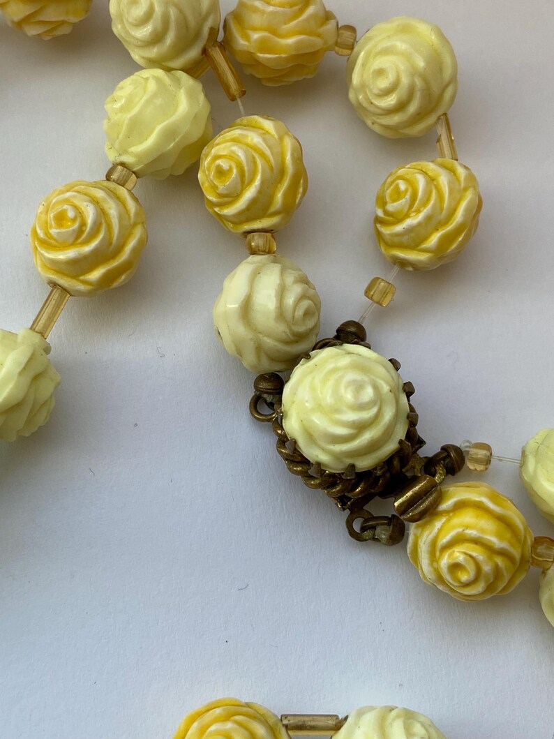 May include: A vintage necklace featuring rose-shaped beads in shades of yellow and cream. The beads are strung together with a delicate chain and clasp. The necklace has a classic, feminine design.