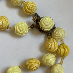 May include: Close-up of a vintage necklace featuring carved rose beads in shades of yellow and cream. The beads are strung together with gold-toned connectors and a decorative clasp. The necklace is a classic piece of jewelry.