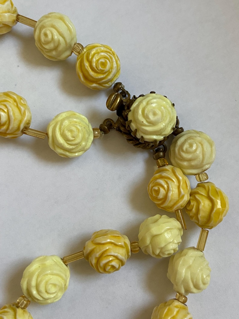 May include: Close-up of a vintage necklace featuring carved rose beads in shades of yellow and cream. The beads are strung together with gold-toned connectors and a decorative clasp. The necklace is a classic piece of jewelry.