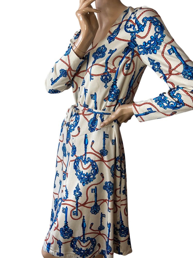 May include: A cream-colored dress with a long sleeve and a V-neck. The dress has a blue and red pattern of keys and rope.