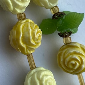 May include: Close-up of a necklace featuring yellow rose-shaped beads and green leaf accents. The beads have a detailed, carved appearance, and are strung together with small, gold-colored spacers. The leaves add a touch of nature to the design.