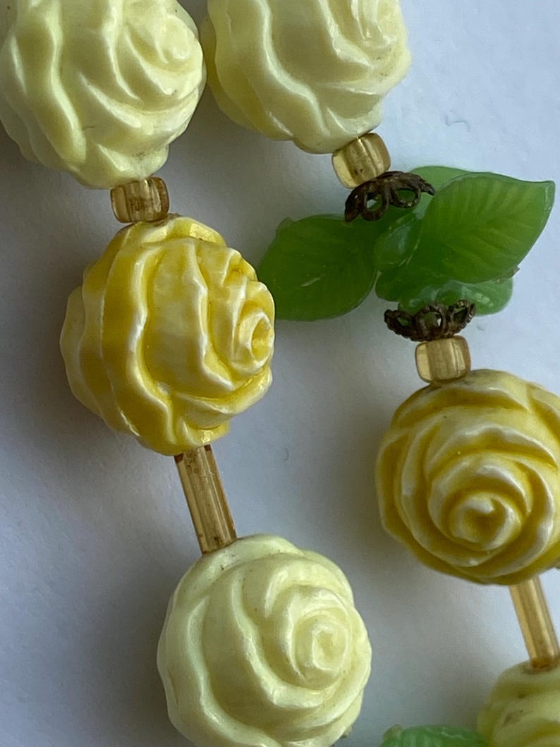 May include: Close-up of a necklace featuring yellow rose-shaped beads and green leaf accents. The beads have a detailed, carved appearance, and are strung together with small, gold-colored spacers. The leaves add a touch of nature to the design.