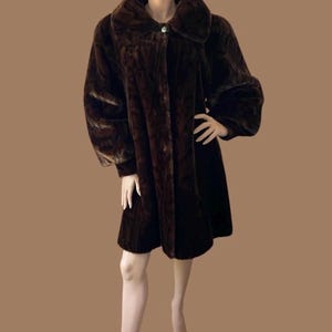 May include: A dark brown, knee-length coat with a plush, textured finish. The coat features a wide, rounded collar, long sleeves with cuffed wrists, and a single visible button closure at the neckline. The garment has a straight, loose fit.