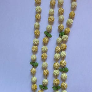 May include: A vintage necklace featuring three strands of cream and yellow rose-shaped beads. Green leaf accents are interspersed throughout the strands. The necklace is secured with a decorative clasp.