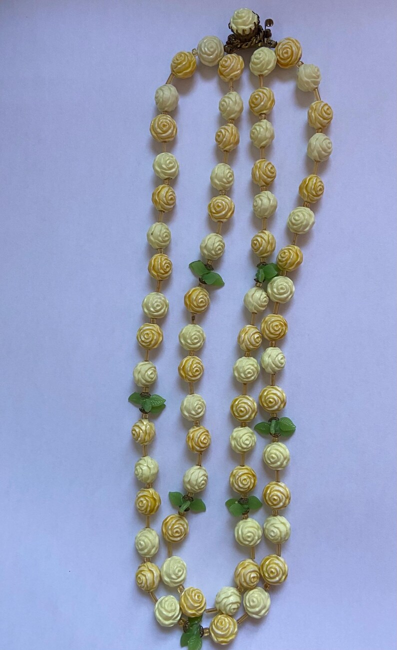 May include: A vintage necklace featuring three strands of cream and yellow rose-shaped beads. Green leaf accents are interspersed throughout the strands. The necklace is secured with a decorative clasp.