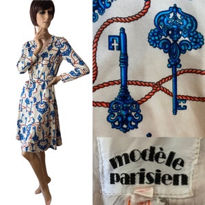 May include: A white dress with a blue and red key and rope pattern. The dress has a V-neckline, long sleeves, and a tie belt. The dress is labeled "mod&egrave;le parisien".