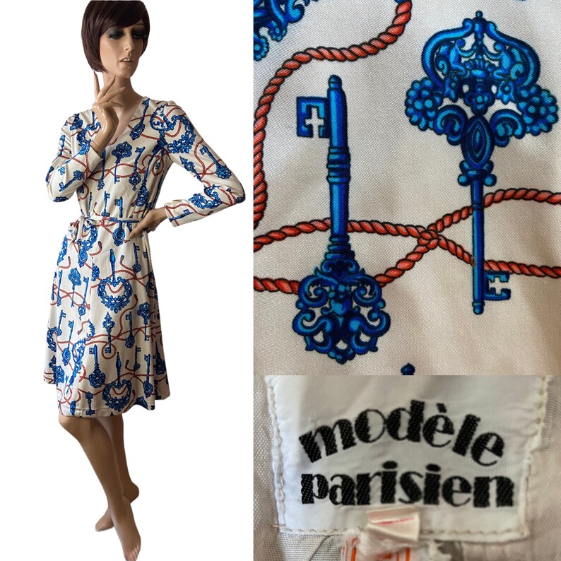 May include: A white dress with a blue and red key and rope pattern. The dress has a V-neckline, long sleeves, and a tie belt. The dress is labeled "mod&egrave;le parisien".
