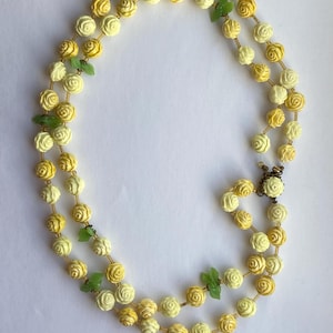 May include: A double-strand necklace featuring cream and yellow rose beads with small green leaf accents. The necklace is secured with a gold-tone clasp. The floral design adds a touch of vintage charm.