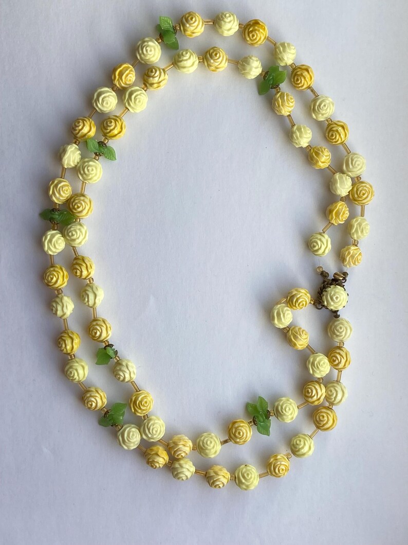 May include: A double-strand necklace featuring cream and yellow rose beads with small green leaf accents. The necklace is secured with a gold-tone clasp. The floral design adds a touch of vintage charm.