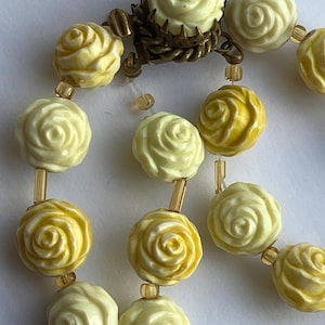 May include: Close-up of a vintage necklace featuring cream and yellow rose-shaped beads. The beads are strung together with gold-toned connectors and a decorative clasp. The necklace has a classic, romantic aesthetic.
