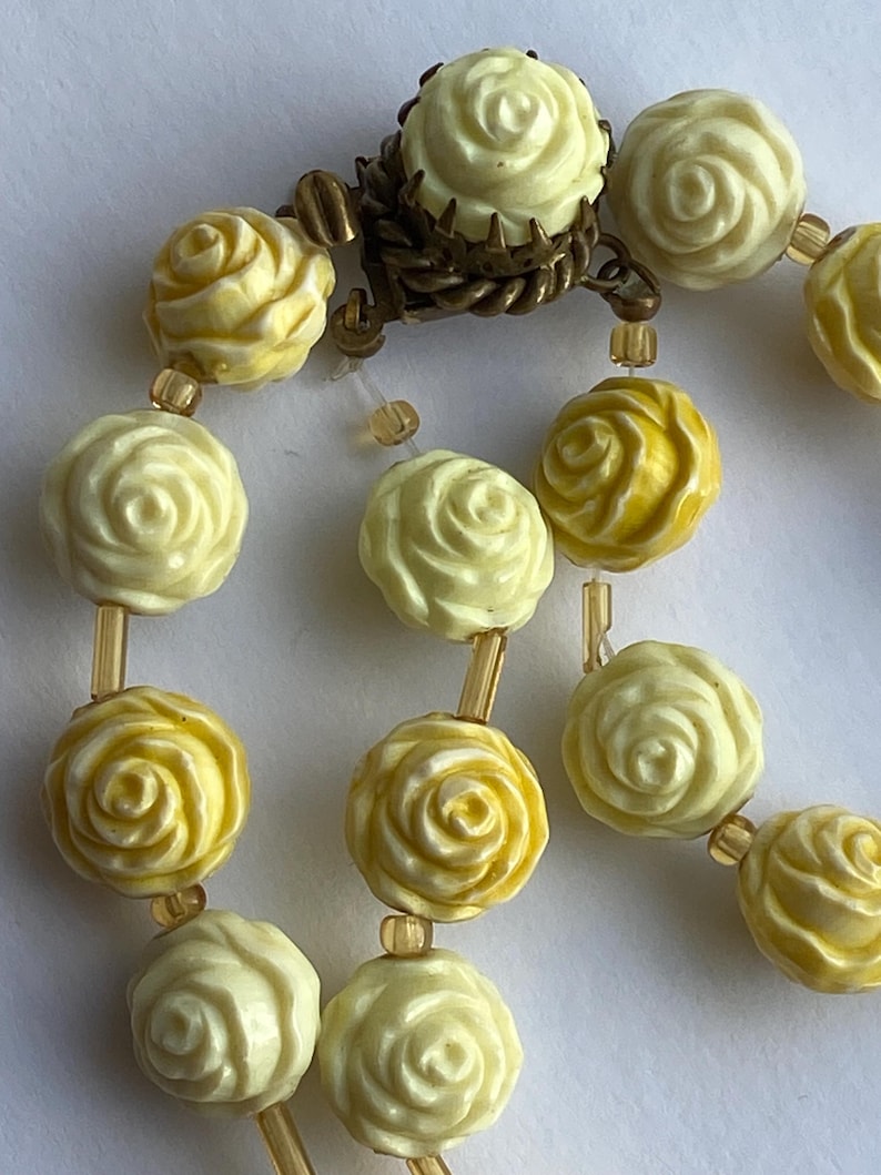 May include: Close-up of a vintage necklace featuring cream and yellow rose-shaped beads. The beads are strung together with gold-toned connectors and a decorative clasp. The necklace has a classic, romantic aesthetic.