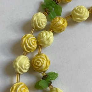 May include: A necklace featuring alternating yellow rose beads and small green leaf accents. The roses vary in shades of yellow, with gold-colored spacers between the beads. The necklace is a vintage style.