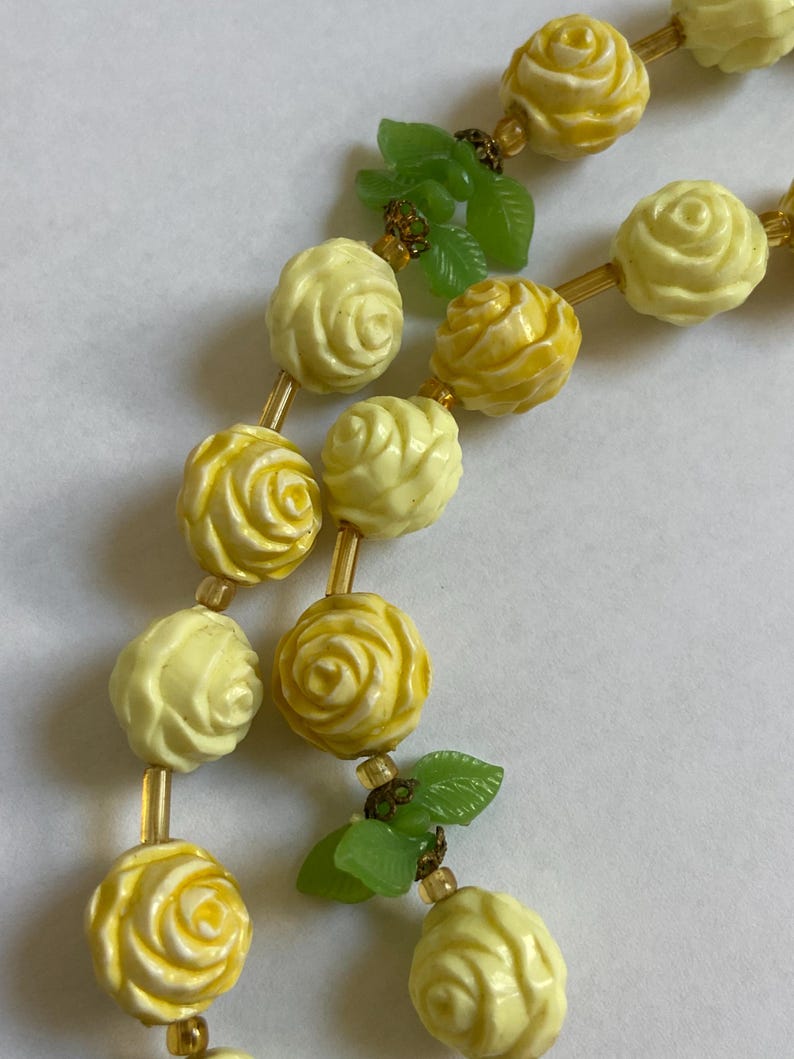 May include: A necklace featuring alternating yellow rose beads and small green leaf accents. The roses vary in shades of yellow, with gold-colored spacers between the beads. The necklace is a vintage style.