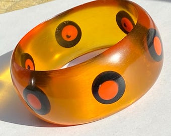 May include: A chunky, translucent orange bangle bracelet with a series of black circles, each containing a smaller orange circle. The bracelet has a smooth, glossy finish and a rounded shape. The design is a bold, retro style.