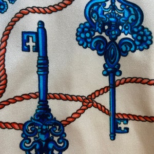 May include: A white fabric with a repeating pattern of blue and orange keys and rope. The keys have intricate designs and are surrounded by a red rope.