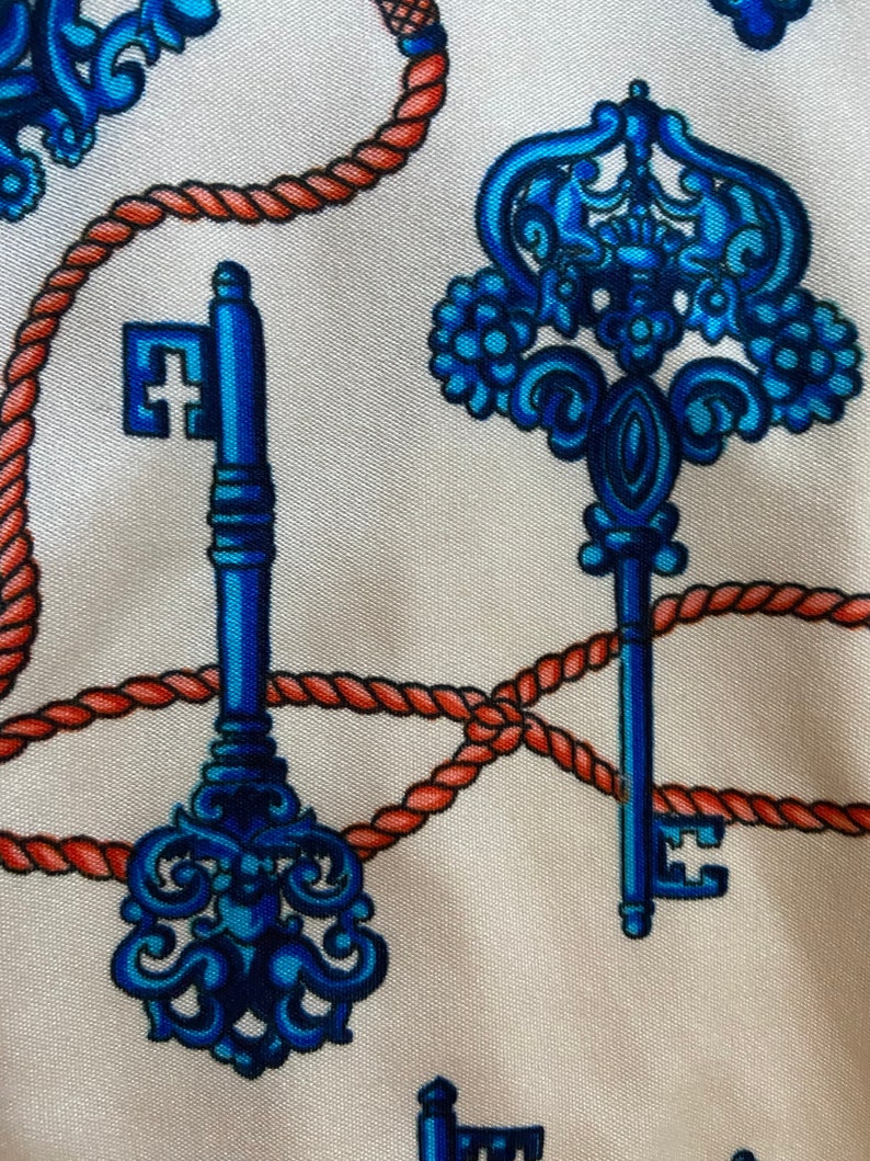 May include: A white fabric with a repeating pattern of blue and orange keys and rope. The keys have intricate designs and are surrounded by a red rope.