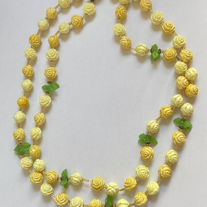 May include: A double-strand necklace featuring cream and yellow rose-shaped beads. The necklace includes small green leaf-shaped accents and a gold-tone clasp. The beads are strung on a delicate gold-colored chain.