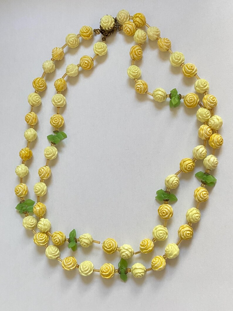 May include: A double-strand necklace featuring cream and yellow rose-shaped beads. The necklace includes small green leaf-shaped accents and a gold-tone clasp. The beads are strung on a delicate gold-colored chain.
