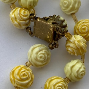 May include: Close-up of a vintage necklace featuring cream and yellow rose-shaped beads. The beads are strung together with small, clear beads and connected by a gold-toned clasp. The necklace has a classic, romantic aesthetic.