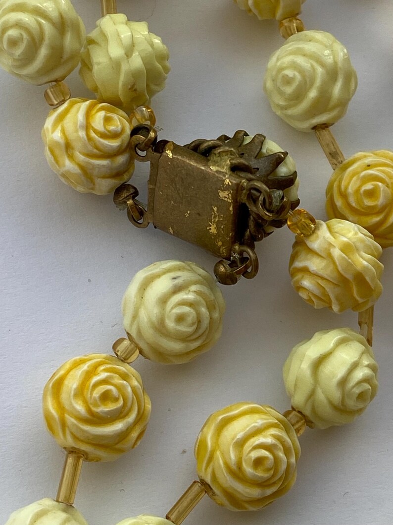 May include: Close-up of a vintage necklace featuring cream and yellow rose-shaped beads. The beads are strung together with small, clear beads and connected by a gold-toned clasp. The necklace has a classic, romantic aesthetic.