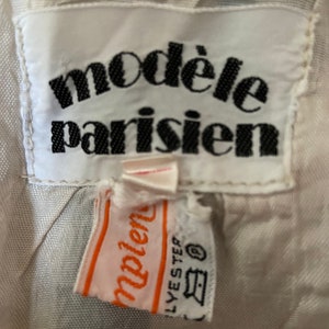 May include: White clothing label with black text that reads "mod&egrave;le parisien". A smaller orange label with white text that reads "mod&egrave;le" is attached to the larger label. The smaller label also has black text that reads "LYESTER" and a symbol that looks like a washing machine.