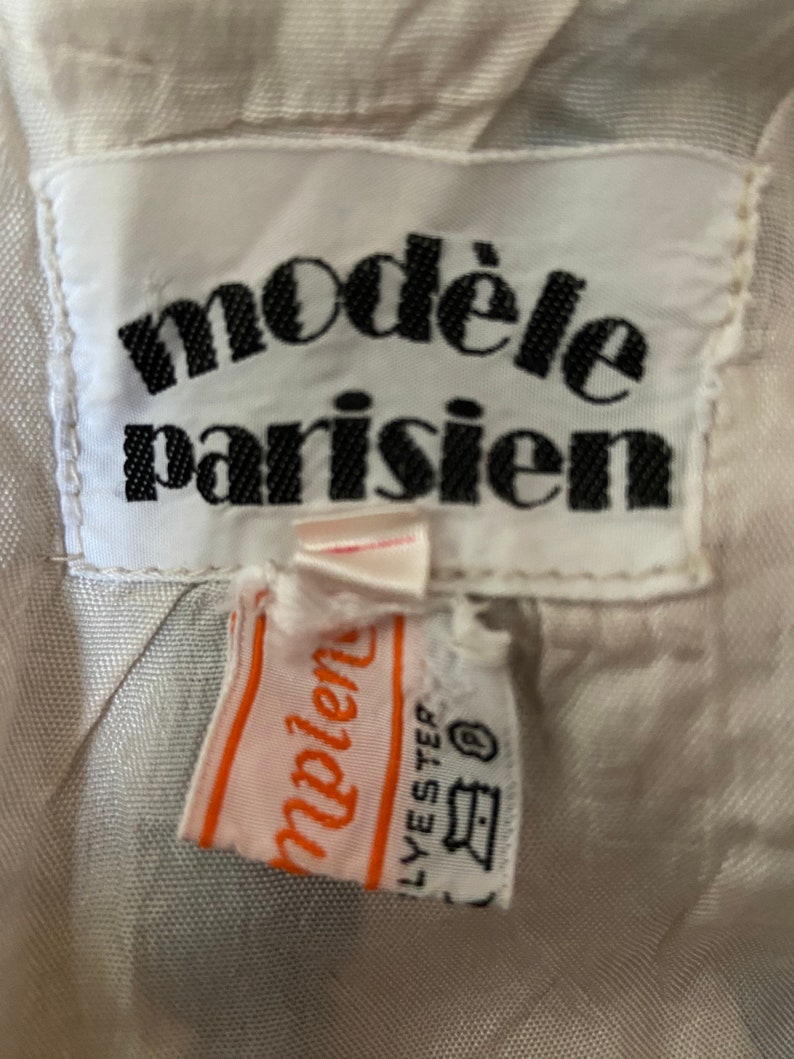 May include: White clothing label with black text that reads "mod&egrave;le parisien". A smaller orange label with white text that reads "mod&egrave;le" is attached to the larger label. The smaller label also has black text that reads "LYESTER" and a symbol that looks like a washing machine.