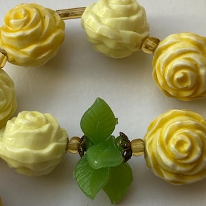 May include: A close-up of a necklace featuring yellow rose-shaped beads and green leaf accents. The beads are connected by gold-colored metal. The necklace is a vintage piece.