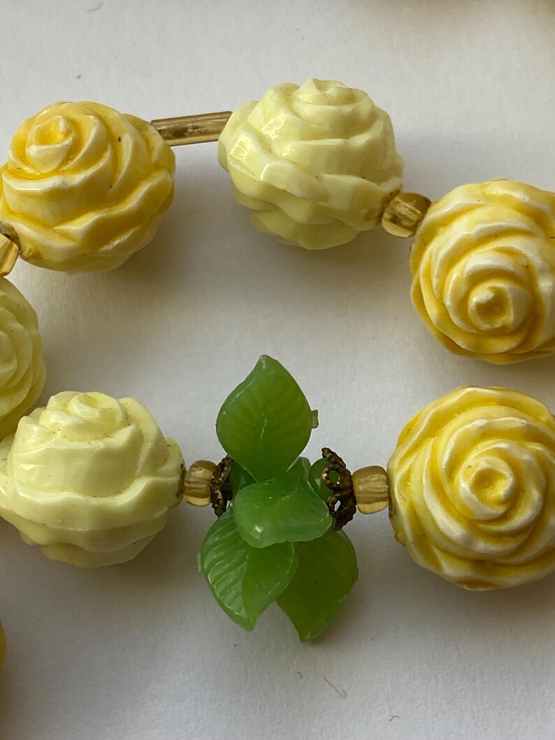 May include: A close-up of a necklace featuring yellow rose-shaped beads and green leaf accents. The beads are connected by gold-colored metal. The necklace is a vintage piece.