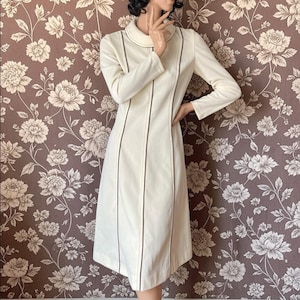 May include: A cream-coloured vintage dress with long sleeves and a collar. The dress has dark brown vertical stripes and trim. The knee-length dress has a tailored fit. The background is a floral wallpaper.